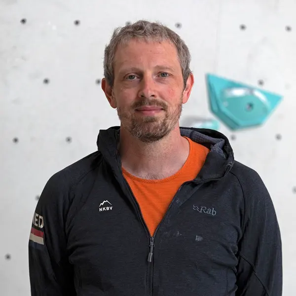 Een man met kort, lichtbruin haar en een baard draagt een oranje shirt en een donker jasje, en staat voor een witte klimwand met een turkoois greepje.